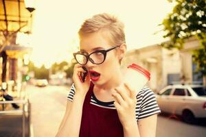 Frau mit Brille reden auf das Telefon auf das Straße ein Tasse von Kaffee foto