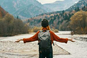 froh Frau Tourist mit ein Rucksack sieht aus beim das Berge in der Nähe von das Fluss auf das Bank foto