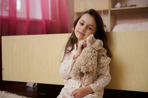 bezaubernd wenig Kind Mädchen im Weiß Pyjama mit Punkte, umarmen ihr Plüsch Spielzeug Schaf und süß lächelnd, suchen beim Kamera foto