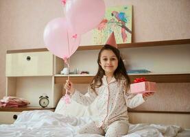 wenig Mädchen im Schlafanzug und Geburtstag Hut, halten Bündel von Rosa Luftballons und Geschenk Kasten, Sitzung auf Bett, lächelnd beim Kamera foto