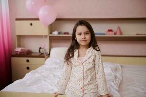 Porträt von selbstbewusst wenig Mädchen mit schön lange Haar, im Pyjama Sitzung auf das Bett im ein Glanz Rosa Schlafzimmer foto