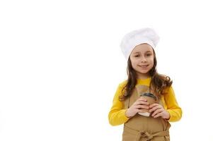 bezaubernd lächelnd wenig Kind Mädchen Kellnerin im Koch Hut und Schürze, halten ein wegbringen heiß trinken im recycelbar Papier Tasse foto