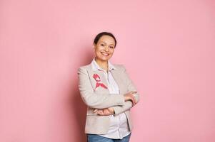 lächelnd positiv Frau tragen Rosa Satin- Band zum zuerst Oktober, posieren mit Waffen gefaltet auf isoliert Rosa Hintergrund foto