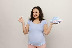 multi ethnisch schwanger Frau halten Neugeborene Kleider im ihr Hand, lächelnd suchen beim Kamera, isoliert auf Weiß Hintergrund foto