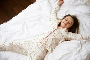 oben Aussicht von bezaubernd wenig Kind Mädchen im Schlafanzug, lächelnd während Lügen Nieder auf ein komfortabel Bett mit orthopädisch Matratze foto