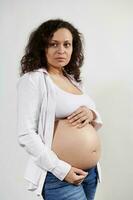 Vertikale Porträt schön schwanger spanisch Frau erwarten ein Baby, posieren mit Hände auf nackt Bauch, Weiß Hintergrund foto