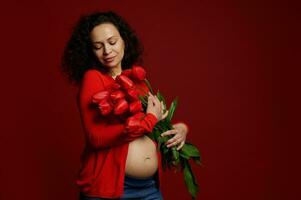 herrlich ziemlich erwartungsvoll Mutter, schwanger Frau mit ein Bündel von rot Tulpen, isoliert auf Farbe Hintergrund. Damen Tag foto