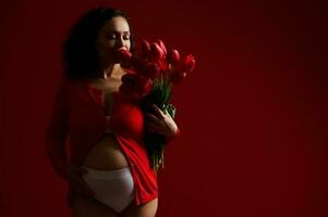 Herrlich Latein amerikanisch Erwachsene schwanger Frau streicheln ihr Bauch, schnüffeln ein süß Strauß von rot Tulpen zum Mutter Tag foto
