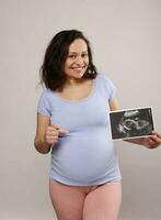 lächelnd schwanger Frau zeigen ihr Neugeborene Baby Sonographie zu das Kamera und zeigen beim ihr Bauch, isoliert auf Weiß foto