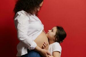 glücklich wenig Mädchen umarmen das Bauch von ihr schwanger Mutter, isoliert auf rot Farbe Hintergrund. glücklich Mutter Tag. Schwangerschaft foto