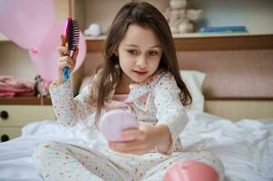 schön Kind Mädchen Sitzung auf ihr Bett, suchen beim ihr Betrachtung im ein Rosa klein Spiegel, nehmen Pflege von ihr lange Haar foto