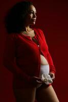 Latein amerikanisch ethnisch Frau im rot Shirt, halten Weiß Orchidee Blume Über ihr schwanger Bauch, suchen verträumt beiseite foto