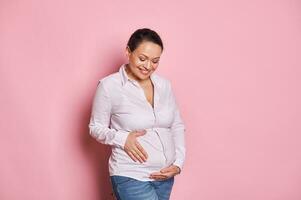 zuversichtlich Porträt von emotional glücklich gravid weiblich lächelnd während streicheln ihr Bauch, isoliert auf Rosa Farbe Hintergrund foto