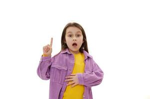 erstaunt aufgeregt wenig Kind Mädchen zeigen Index Finger beiseite hoch, suchen beim Kamera mit ihr Mund offen, Weiß Hintergrund foto
