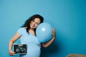 glücklich schwanger Frau halten Blau Ballon und Ultraschall Scan von ihr Zukunft Baby Junge, isoliert Hintergrund. Geschlecht Party foto