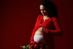 Mode Porträt Schuss im niedrig Schlüssel von ein glücklich schwanger Frau streicheln ihr Bauch, lächelnd posieren mit ein Strauß von Tulpen foto