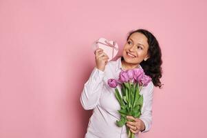 glücklich schwanger Frau mit wellig Haar, halten Strauß von Tulpen, lächelnd, zittern Geschenk Kasten, raten was ist Innerhalb es foto