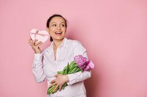 attraktiv Frau mit Strauß von Tulpen, zittern ein Geschenk Kasten, versuchen zu vermuten Was ist Innerhalb Es, isoliert Rosa Hintergrund foto