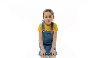 kaukasisch wenig Mädchen im Gelb T-Shirt und Denim Sommerkleid halten Hände auf ihr Knie während Sitzung auf Weiß Hintergrund foto