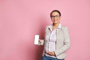 Mitte östlichen schwanger Frau im Brille, berührt Bauch, lächelt, zeigt an Zelle Telefon mit Weiß Digital Bildschirm. Attrappe, Lehrmodell, Simulation foto