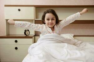bezaubernd Baby Mädchen im stilvoll Pyjama, aufwachen oben Dehnen im das Morgen im ihr Schlafzimmer beim Zuhause foto