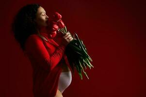 Seite Porträt auf rot Hintergrund von schön schwanger Frau schnüffeln ein Strauß von rot Tulpen. glücklich Mutter Tag Konzept foto