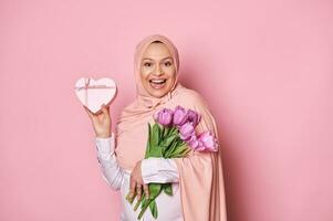 Muslim schwanger Frau im Rosa Kopftuch, halten ein Strauß von lila Tulpen und ein Herz geformt Geschenk Box zum Mutter Tag foto