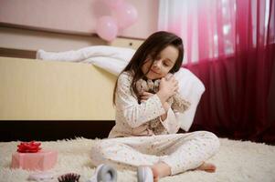 herrlich 5-6 Jahre alt Kind Mädchen im Pyjama, Sitzung auf das Teppich im ihr kindisch Zimmer und umarmen ihr Plüsch Spielzeug foto