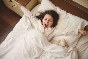 Aussicht von über von bezaubernd wenig Kind Mädchen Ausstrecken Waffen während aufwachen oben im das Morgen im glücklich Stimmung foto