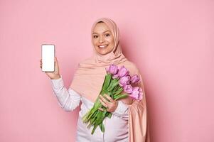 lächelnd schwanger Muslim Frau im Rosa Kopftuch, lächelt zeigt an beim Kamera ein Smartphone mit Weiß leer Bildschirm mit Kopieren Raum foto