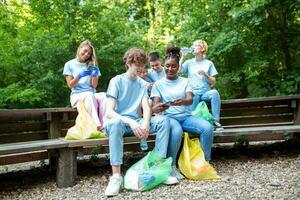 spontan Bild von ein klein Gruppe von Freiwillige mit Handschuhe und Müll Taschen Herstellung ein halt während im das Park Sie haben gerade gereinigt. alle tragen Blau T-Shirts, Sein stolz und glücklich zum Was Sie tat foto