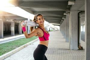 sportliche junge Frau mit Handtuch lächelnd und streckend. junge sportliche frauenausbildung. frau, die trainingsübungen auf der straße macht. fittes Mädchen, das sich vor dem aktiven Fitnesstraining ausbreitet foto