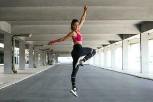 Fitness Frau Springen draussen im städtisch Umfeld. aktiv Frau Sein voll von Energie, springt hoch im Luft, bereitet vor zum Sport Wettbewerbe. . Gymnastik Konzept foto