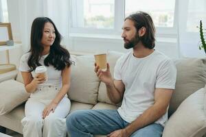 Mann und Frau Sitzung beim Zuhause auf das Couch im Weiß stilvoll T-Shirts Trinken Kaffee aus von Krabbe Tassen von ein Kaffee Geschäft und haben Spaß chatten lächelt und Lachen beim heim. männlich und weiblich Freundschaft foto