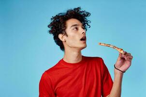 Mann mit ein Stück von Pizza im seine Hand auf ein Blau Hintergrund Teenager im ein rot T-Shirt lockig Haar foto