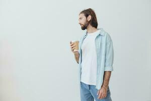 freiberuflich tausendjährig Mann mit Bart Trinken Kaffee von ein Krabbe Tasse im stilvoll Hipster Kleider auf ein Weiß Hintergrund foto