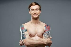 sexy Bodybuilder mit nackt Torso gemacht von Muskel Tätowierungen grau Hintergrund gekreuzt Waffen auf Truhe foto