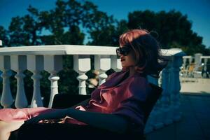 glamourös Frau im Sonnenbrille draußen Sitzung auf das Terrasse entspannend sonnig Tag foto