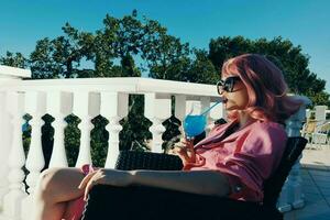 glamourös Frau im Sonnenbrille draußen Sitzung auf das Terrasse Cocktail entspannend Lebensstil foto