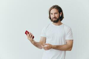 ein Mann mit ein Hipster Bart im ein Weiß T-Shirt mit ein Telefon und kabellos Kopfhörer lächelnd Hören zu Musik- und ein Hörbuch online gegen ein Weiß Mauer foto