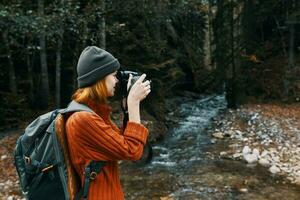 Frau Fotograf halten ein Kamera im Hand in der Nähe von das Fluss im das Berge und Wald im das Hintergrund foto