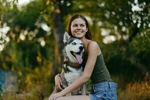 froh Frau mit ein heiser Rasse Hund lächelt während Sitzung im Natur auf ein gehen mit ein Hund auf ein Leine Herbst Landschaft auf das Hintergrund. Lebensstil im Spaziergänge mit Haustiere foto