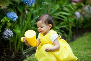 Süss einer Jahr alt Baby Mädchen gekleidet im Gelb Bewässerung das Pflanzen beim das Garten foto