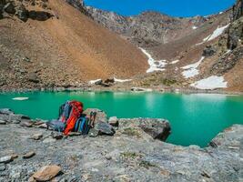 ein groß Orange Rucksack und Trekking Stangen auf das Ufer von ein Berg See. Wandern im das Hochland. das Zeit von das halt auf das Wanderung. foto