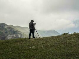 Fotograf schießt ein Landschaft mit ein Stativ während Stehen auf das Kante von ein Cliff im schwierig Wetter Bedingungen. foto
