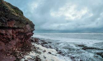 ein Felsen Cliff über das Wasser mit ein Gezeiten Küstenlinie. Weiß Meer. foto