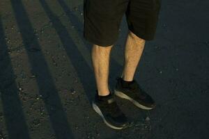 Beine von Kerl im schwarz kurze Hose und schwarz Turnschuhe. Personen Füße Stand auf Arsch. kurze Hose im Mode. foto