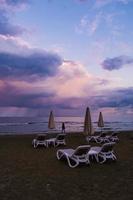 Die Silhouette einer Frau wird während der blauen Stunde an einem stürmischen Tag am McKenzie Beach, Larnaca, Zypern gegen das Meer, die Sonnenschirme und die Sonnenliegen gesetzt foto