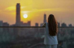 Frau, die den Blick auf den Sonnenuntergang vom Balkon aus betrachtet und genießt, während die Sonne hinter dem Wolkenkratzer in der geschäftigen Innenstadt untergeht, mit Einsamkeit für Einsamkeit, Einsamkeit und Träumen vom Lebensstil der Freiheit foto