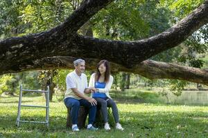 asiatisch Senior Vater mit Gehhilfe und Tochter Sitzung zusammen unter das groß Baum im das Park während Sommer- zum Licht Übung und physisch Therapie Verwendung. foto
